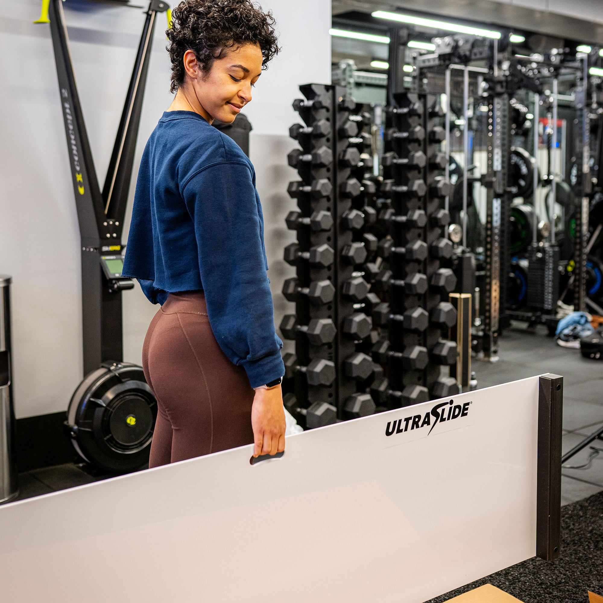 Person carrying an UltraSlide slide board with its built-in handle in a gym setting with dumbbells and exercise equipment in the background