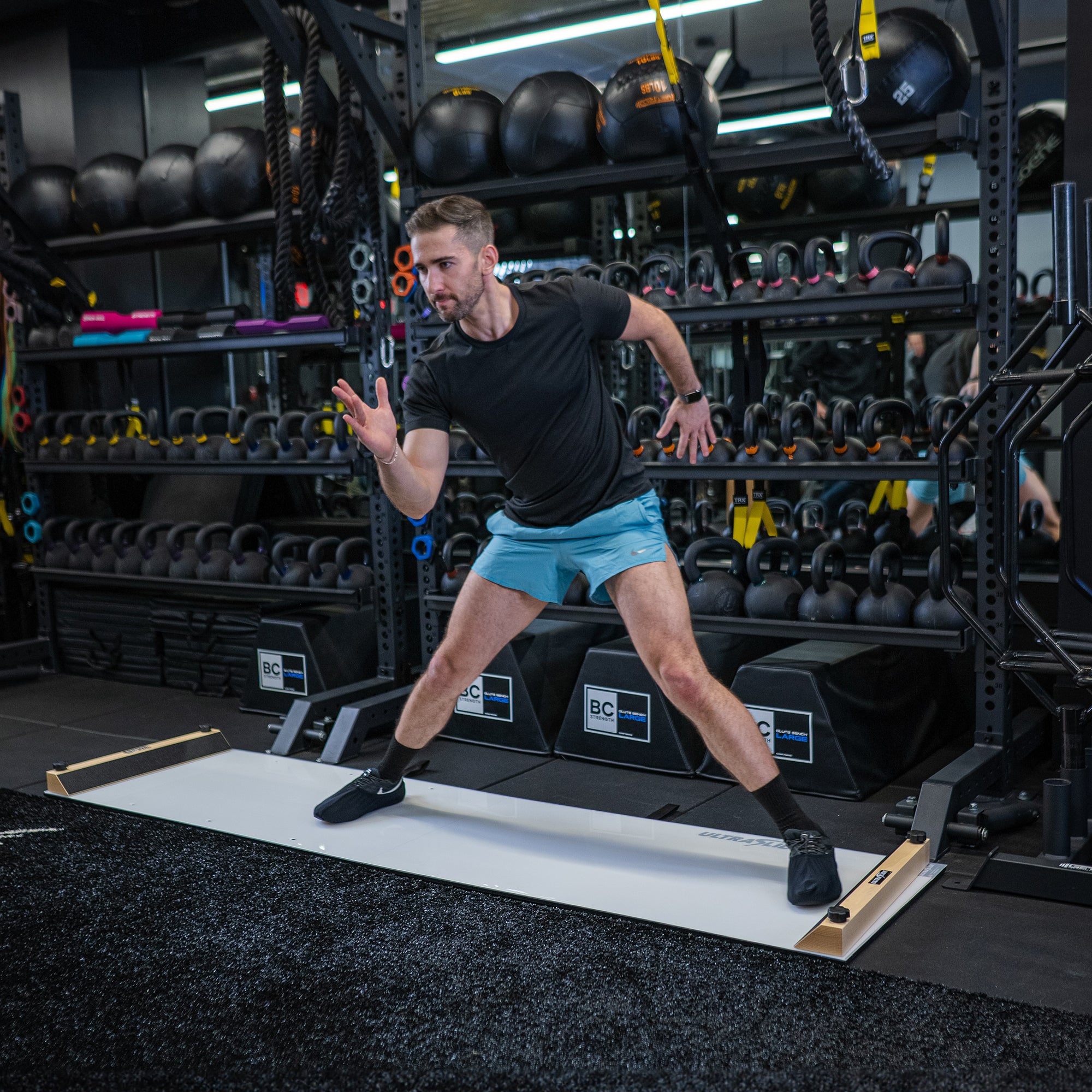 Man using an ULTRASLIDE slide board in a gym setting with equipment in the background