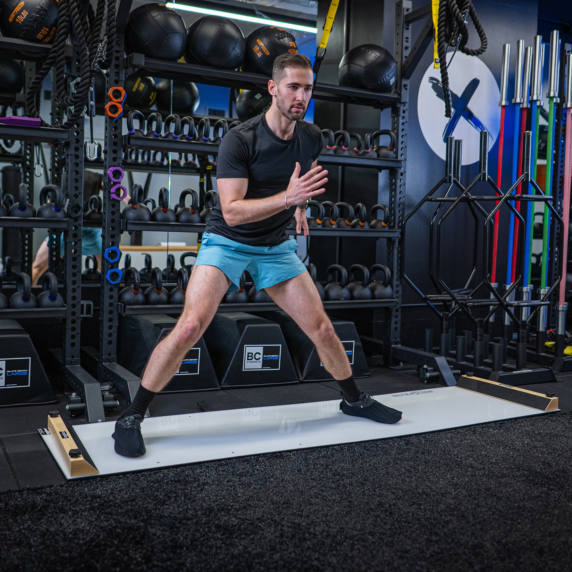 Man exercising on an ULTRASLIDE slide board in a gym setting with equipment and weights in the background