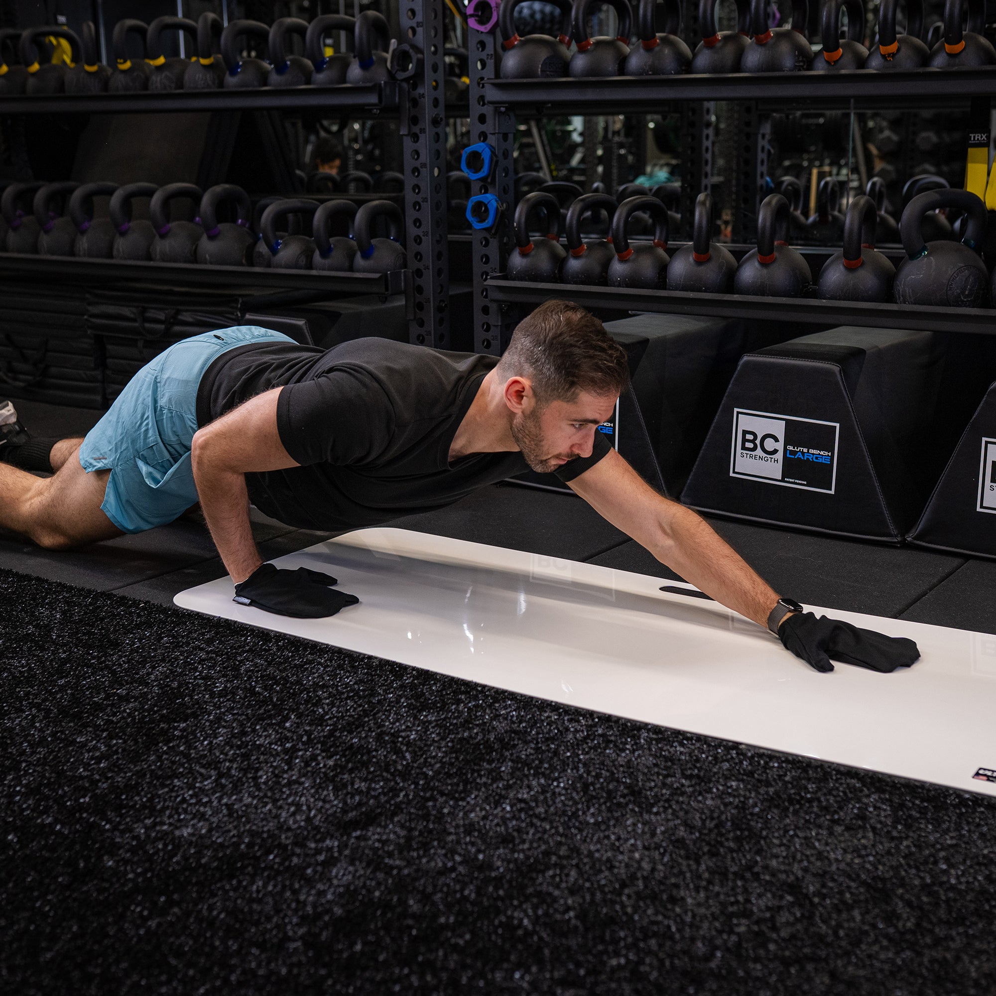Man wearing ULTRASLIDE slide board mitts while working out on a slide board in a gym setting with weight racks in the background.